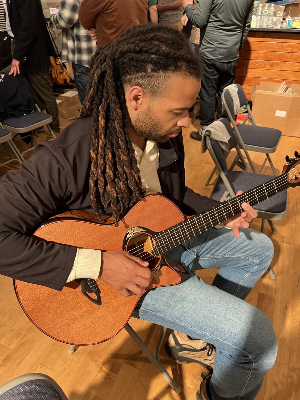 A guitarist seated in the room playing an acoustic guitar while other attendees talk nearby.